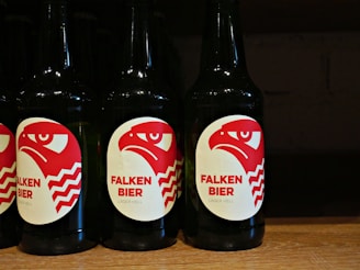 Several dark-colored beer bottles are displayed with labels featuring a red and white stylized eagle head and the text 'FALKEN BIER' along with 'LAGER HELL' printed on them. The bottles are neatly arranged in a row on a wooden shelf.