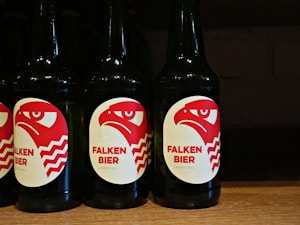 Several dark-colored beer bottles are displayed with labels featuring a red and white stylized eagle head and the text 'FALKEN BIER' along with 'LAGER HELL' printed on them. The bottles are neatly arranged in a row on a wooden shelf.
