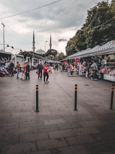 A bustling outdoor marketplace scene with various people walking and interacting. Stalls line the right side, displaying clothing and other goods. In the background, two tall minarets rise into a cloudy sky, suggesting the proximity of a mosque. Trees border the right side, and streetlights and cables are visible overhead.