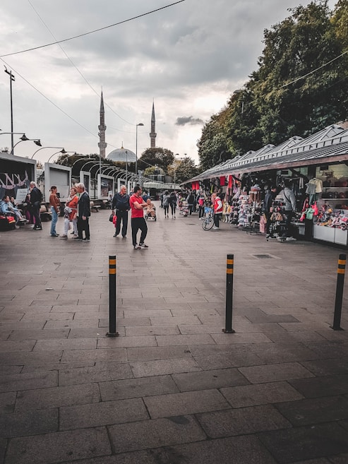 A bustling outdoor marketplace scene with various people walking and interacting. Stalls line the right side, displaying clothing and other goods. In the background, two tall minarets rise into a cloudy sky, suggesting the proximity of a mosque. Trees border the right side, and streetlights and cables are visible overhead.