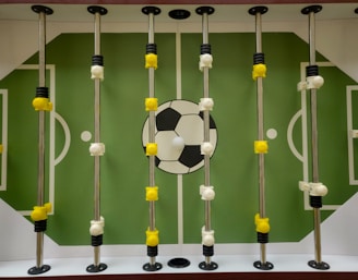 A foosball table with a green playing field and white markings resembling a soccer field. Rotating metal rods are placed across the field, with yellow and white foosball player figures attached to these rods. A large soccer ball graphic is located at the center of the field.