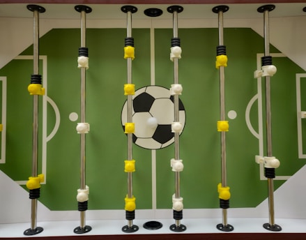 A foosball table with a green playing field and white markings resembling a soccer field. Rotating metal rods are placed across the field, with yellow and white foosball player figures attached to these rods. A large soccer ball graphic is located at the center of the field.