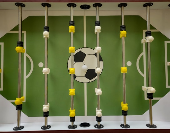 A foosball table with a green playing field and white markings resembling a soccer field. Rotating metal rods are placed across the field, with yellow and white foosball player figures attached to these rods. A large soccer ball graphic is located at the center of the field.