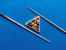 A set of billiard balls arranged in a triangular rack on a bright blue pool table surface, flanked by two pool cues placed diagonally.