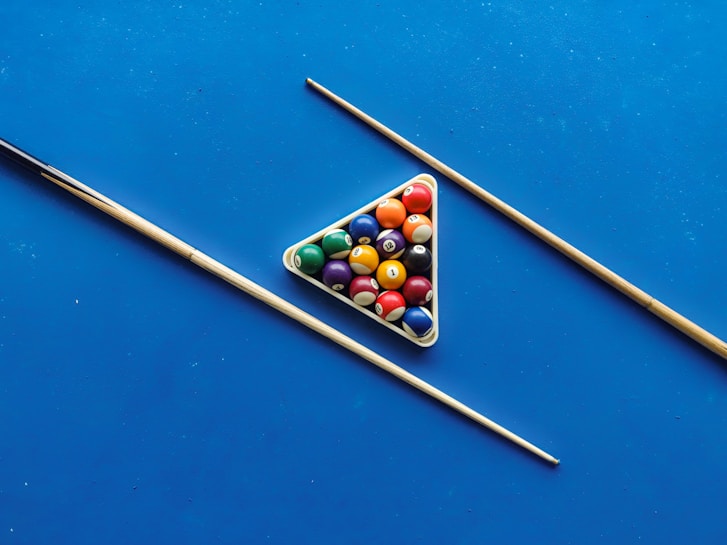A set of billiard balls arranged in a triangular rack on a bright blue pool table surface, flanked by two pool cues placed diagonally.