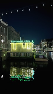A nighttime scene featuring a canal with a small illuminated building labeled 'Rondvaart Koou'. The building is lighted with bright green lights, reflecting off the water. A boat is moored next to the building with the name 'Reederij Boeckl' visible on it. Surrounding architecture includes traditional canal-side buildings.