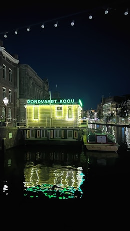 A nighttime scene featuring a canal with a small illuminated building labeled 'Rondvaart Koou'. The building is lighted with bright green lights, reflecting off the water. A boat is moored next to the building with the name 'Reederij Boeckl' visible on it. Surrounding architecture includes traditional canal-side buildings.