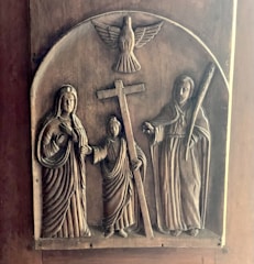 A carved wooden relief depicting three robed figures, one holding a cross, with a dove above them. The figures appear to be religious, with two on either side of the cross. The craftsmanship highlights intricate details in the robes and facial expressions.