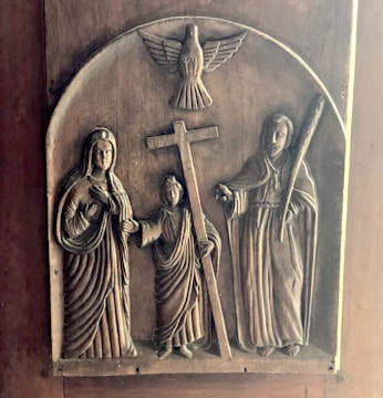 A carved wooden relief depicting three robed figures, one holding a cross, with a dove above them. The figures appear to be religious, with two on either side of the cross. The craftsmanship highlights intricate details in the robes and facial expressions.
