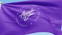 A vibrant purple flag with the words 'Agenda Edu' prominently displayed in the center in a cursive font. Surrounding the central text are the words 'Simplificamos Jornadas Educacionais' arranged in a circular pattern. The flag waves gently, creating ripples in the fabric against a clear blue sky.