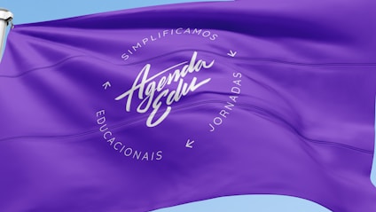 A vibrant purple flag with the words 'Agenda Edu' prominently displayed in the center in a cursive font. Surrounding the central text are the words 'Simplificamos Jornadas Educacionais' arranged in a circular pattern. The flag waves gently, creating ripples in the fabric against a clear blue sky.