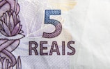 A close-up view of a Brazilian 5 reais banknote featuring intricate designs and patterns. The number '5' is prominently displayed in blue, with the word 'REAIS' below it in a stylized font. There are floral and geometric motifs in the background.