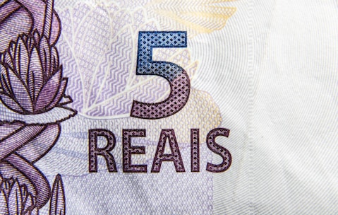 A close-up view of a Brazilian 5 reais banknote featuring intricate designs and patterns. The number '5' is prominently displayed in blue, with the word 'REAIS' below it in a stylized font. There are floral and geometric motifs in the background.