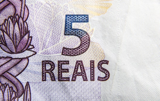 A close-up view of a Brazilian 5 reais banknote featuring intricate designs and patterns. The number '5' is prominently displayed in blue, with the word 'REAIS' below it in a stylized font. There are floral and geometric motifs in the background.