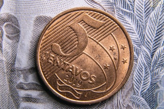 A close-up of a 5 centavos coin from 2019 resting on a banknote with intricate line patterns. The coin features engraved lines, stars, and the denomination.