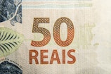 Close-up of a 50 reais banknote featuring intricate patterns and textures. The denomination '50' is prominently displayed in a reddish-brown color with the word 'REAIS' beneath it. Decorative elements and fine detailing can be seen throughout the design.