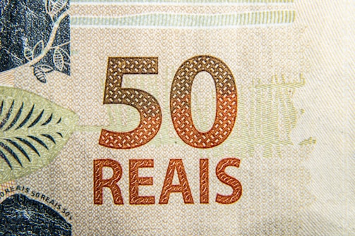 Close-up of a 50 reais banknote featuring intricate patterns and textures. The denomination '50' is prominently displayed in a reddish-brown color with the word 'REAIS' beneath it. Decorative elements and fine detailing can be seen throughout the design.
