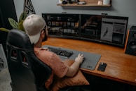 A person wearing a cap is seated at a desk with dual monitors displaying digital content. The workspace includes a large desk mat with a keyboard and mouse, and a smartphone is placed on the desk. A gaming chair provides comfort, and there are decorative items like a model car and plants on a shelf above the monitors.