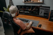 A person wearing a cap is seated at a desk with dual monitors displaying digital content. The workspace includes a large desk mat with a keyboard and mouse, and a smartphone is placed on the desk. A gaming chair provides comfort, and there are decorative items like a model car and plants on a shelf above the monitors.
