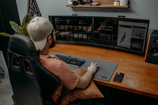 A friendly Muslim gamer at a desk, smiling while typing on a laptop surrounded by gaming gear.