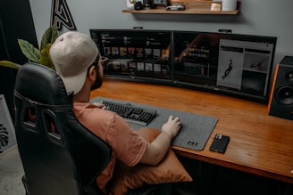 A person wearing a cap is seated at a desk with dual monitors displaying digital content. The workspace includes a large desk mat with a keyboard and mouse, and a smartphone is placed on the desk. A gaming chair provides comfort, and there are decorative items like a model car and plants on a shelf above the monitors.