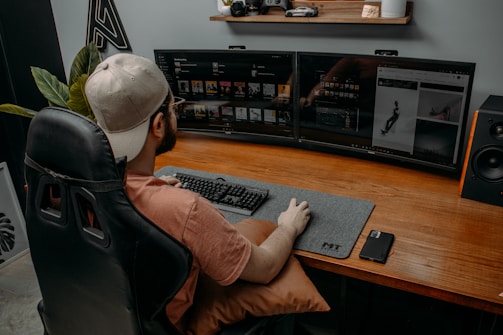A person wearing a cap is seated at a desk with dual monitors displaying digital content. The workspace includes a large desk mat with a keyboard and mouse, and a smartphone is placed on the desk. A gaming chair provides comfort, and there are decorative items like a model car and plants on a shelf above the monitors.