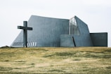 A modern, angular church building with concrete walls and large windows is situated on a grassy field. A large cross is positioned to the left of the structure, adding a religious significance to the scene.