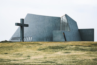 A modern, angular church building with concrete walls and large windows is situated on a grassy field. A large cross is positioned to the left of the structure, adding a religious significance to the scene.