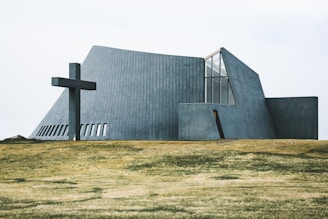 A modern, angular church building with concrete walls and large windows is situated on a grassy field. A large cross is positioned to the left of the structure, adding a religious significance to the scene.