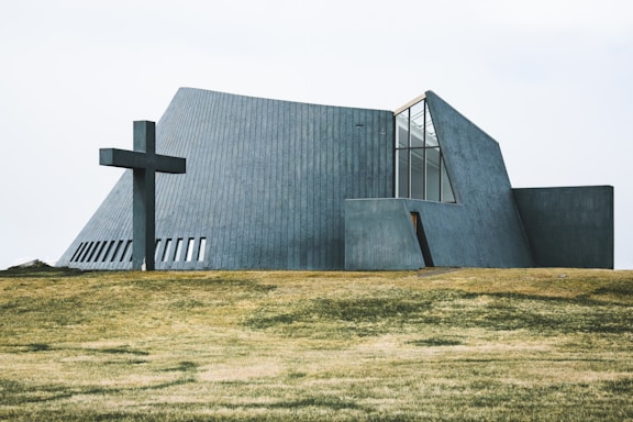 A modern, angular church building with concrete walls and large windows is situated on a grassy field. A large cross is positioned to the left of the structure, adding a religious significance to the scene.