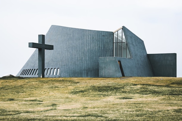 A modern, angular church building with concrete walls and large windows is situated on a grassy field. A large cross is positioned to the left of the structure, adding a religious significance to the scene.