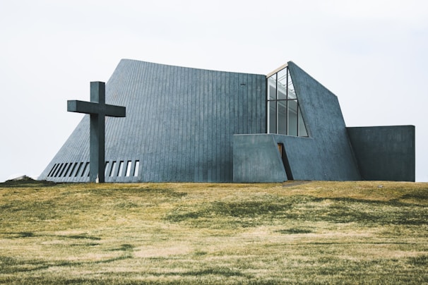 A modern, angular church building with concrete walls and large windows is situated on a grassy field. A large cross is positioned to the left of the structure, adding a religious significance to the scene.