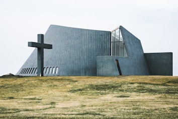 A modern, angular church building with concrete walls and large windows is situated on a grassy field. A large cross is positioned to the left of the structure, adding a religious significance to the scene.