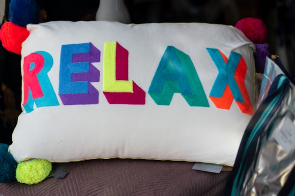 Image of white cushion with word RELAX in bright colours on it. Pop moms on the each corner of the cushion.