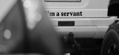 A black and white photograph of the rear of a vehicle with an out-of-focus foreground. The vehicle's prominent feature is the text 'I'm a servant' displayed on its back. The image includes visible vehicle accessories like a tow hitch and part of a tire.
