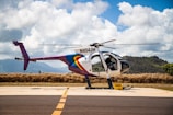 A helicopter with registration N454JH is parked on a tarmac against a backdrop of lush hills and a partly cloudy sky. The helicopter features a colorful design, with prominent rainbow-like stripes on the fuselage. Some aviation equipment and a fuel container are visible nearby.