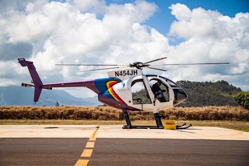 A helicopter with registration N454JH is parked on a tarmac against a backdrop of lush hills and a partly cloudy sky. The helicopter features a colorful design, with prominent rainbow-like stripes on the fuselage. Some aviation equipment and a fuel container are visible nearby.