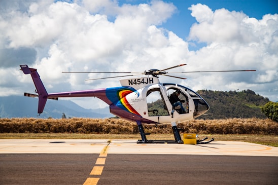 A helicopter with registration N454JH is parked on a tarmac against a backdrop of lush hills and a partly cloudy sky. The helicopter features a colorful design, with prominent rainbow-like stripes on the fuselage. Some aviation equipment and a fuel container are visible nearby.