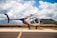 A helicopter with registration N454JH is parked on a tarmac against a backdrop of lush hills and a partly cloudy sky. The helicopter features a colorful design, with prominent rainbow-like stripes on the fuselage. Some aviation equipment and a fuel container are visible nearby.