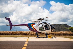 A helicopter with registration N454JH is parked on a tarmac against a backdrop of lush hills and a partly cloudy sky. The helicopter features a colorful design, with prominent rainbow-like stripes on the fuselage. Some aviation equipment and a fuel container are visible nearby.