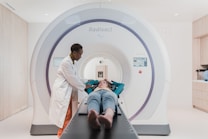 A person in a lab coat stands beside a patient who is lying down on a table that is part of a large medical imaging machine. The setting appears to be a clean and modern medical facility.