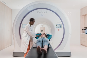 A person in a lab coat stands beside a patient who is lying down on a table that is part of a large medical imaging machine. The setting appears to be a clean and modern medical facility.