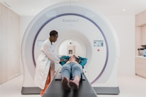 A person in a lab coat stands beside a patient who is lying down on a table that is part of a large medical imaging machine. The setting appears to be a clean and modern medical facility.
