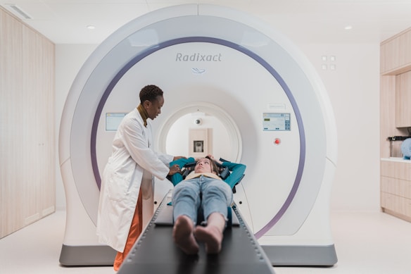 A person in a lab coat stands beside a patient who is lying down on a table that is part of a large medical imaging machine. The setting appears to be a clean and modern medical facility.