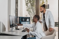 Two people in lab coats collaborate in an office setting. One is seated and pointing at a computer screen displaying medical images, while the other stands nearby holding a tablet. Large windows reveal a view of greenery outside, creating a bright and professional environment.