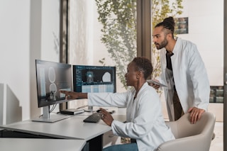 Two people in lab coats collaborate in an office setting. One is seated and pointing at a computer screen displaying medical images, while the other stands nearby holding a tablet. Large windows reveal a view of greenery outside, creating a bright and professional environment.