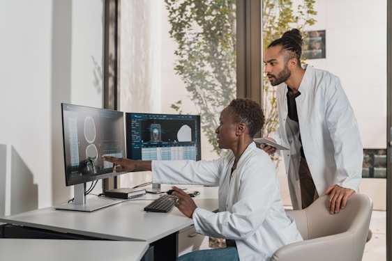 Two people in lab coats collaborate in an office setting. One is seated and pointing at a computer screen displaying medical images, while the other stands nearby holding a tablet. Large windows reveal a view of greenery outside, creating a bright and professional environment.