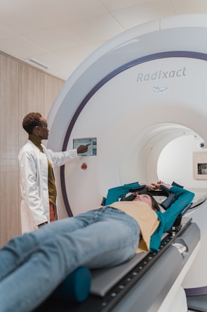 A person in a medical setting is lying on a table, about to enter a large cylindrical medical imaging machine labeled 'Radixact'. Another person, wearing a white coat, stands beside the machine, using a touchscreen panel.