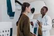 Two women are engaged in conversation in a medical setting, one wearing a white coat, suggesting she is a healthcare professional. The environment contains medical equipment, possibly indicating a check-up or consultation is taking place.