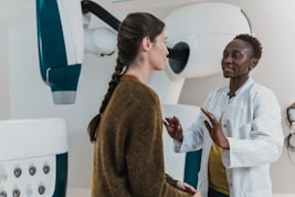 Two women are engaged in conversation in a medical setting, one wearing a white coat, suggesting she is a healthcare professional. The environment contains medical equipment, possibly indicating a check-up or consultation is taking place.